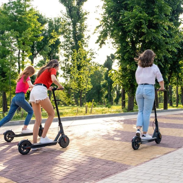E-scooter, electric scooter, Ecological urban city transport, alternative energy, alternative lifestyle. Three Young girl friends on vacation having fun driving electric scooter through city park.